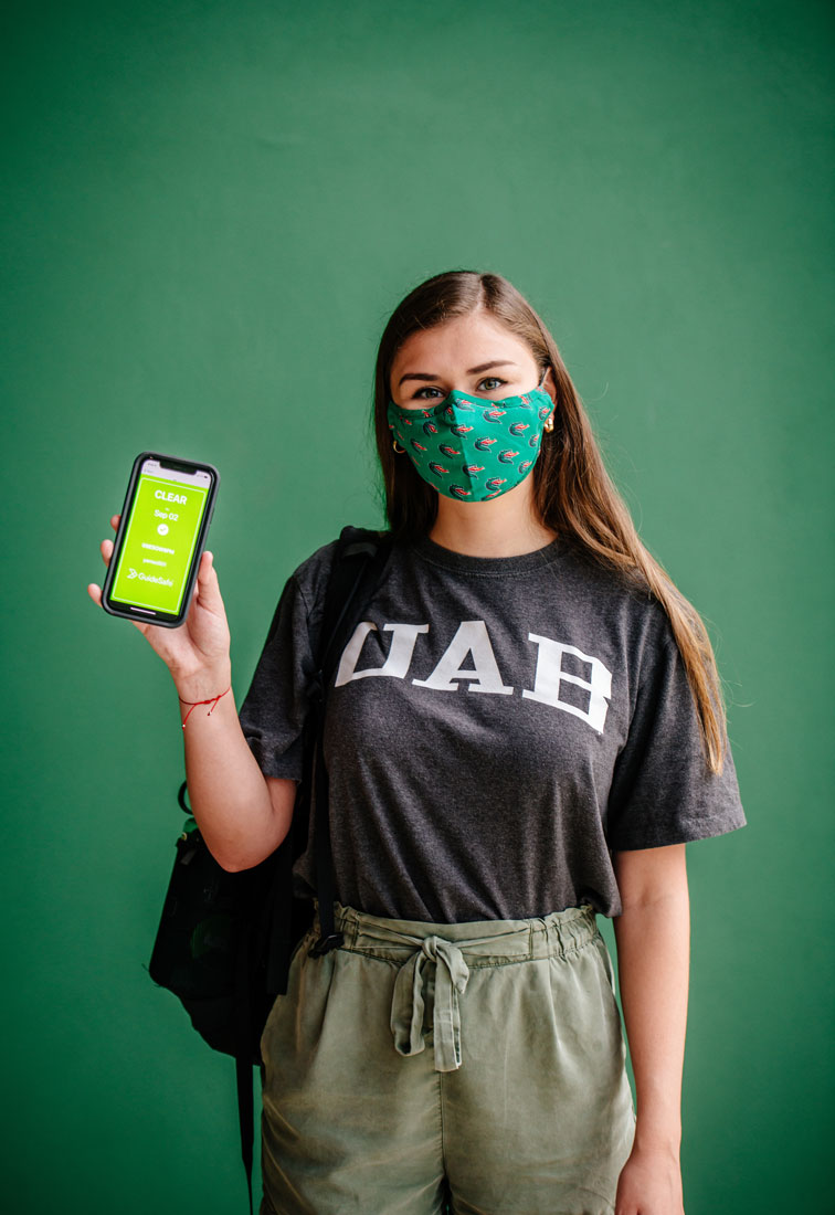 A young student wearing a UAB mask is holding a phone with the COVID-19 tracker app open.