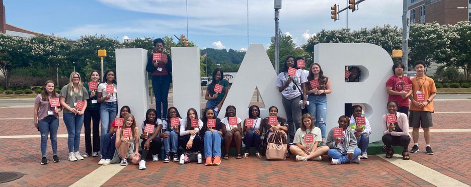 Summer institute students pose around and in front of giant white cut-out UAB letters on brick sidewalk.