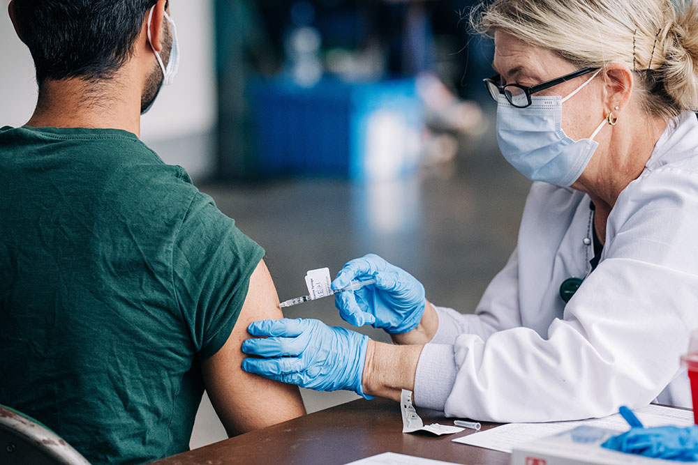 Back, a male student is getting vaccinated against COVID-19 (Coronavirus Disease) by a health care worker wearing a face mask at Bartow Arena, May 18, 2021.
