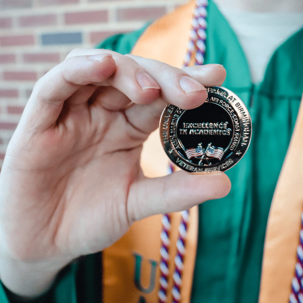 Student model wearing graduation gown and Veterans Services honor cord, holding Veterans Services honors coin