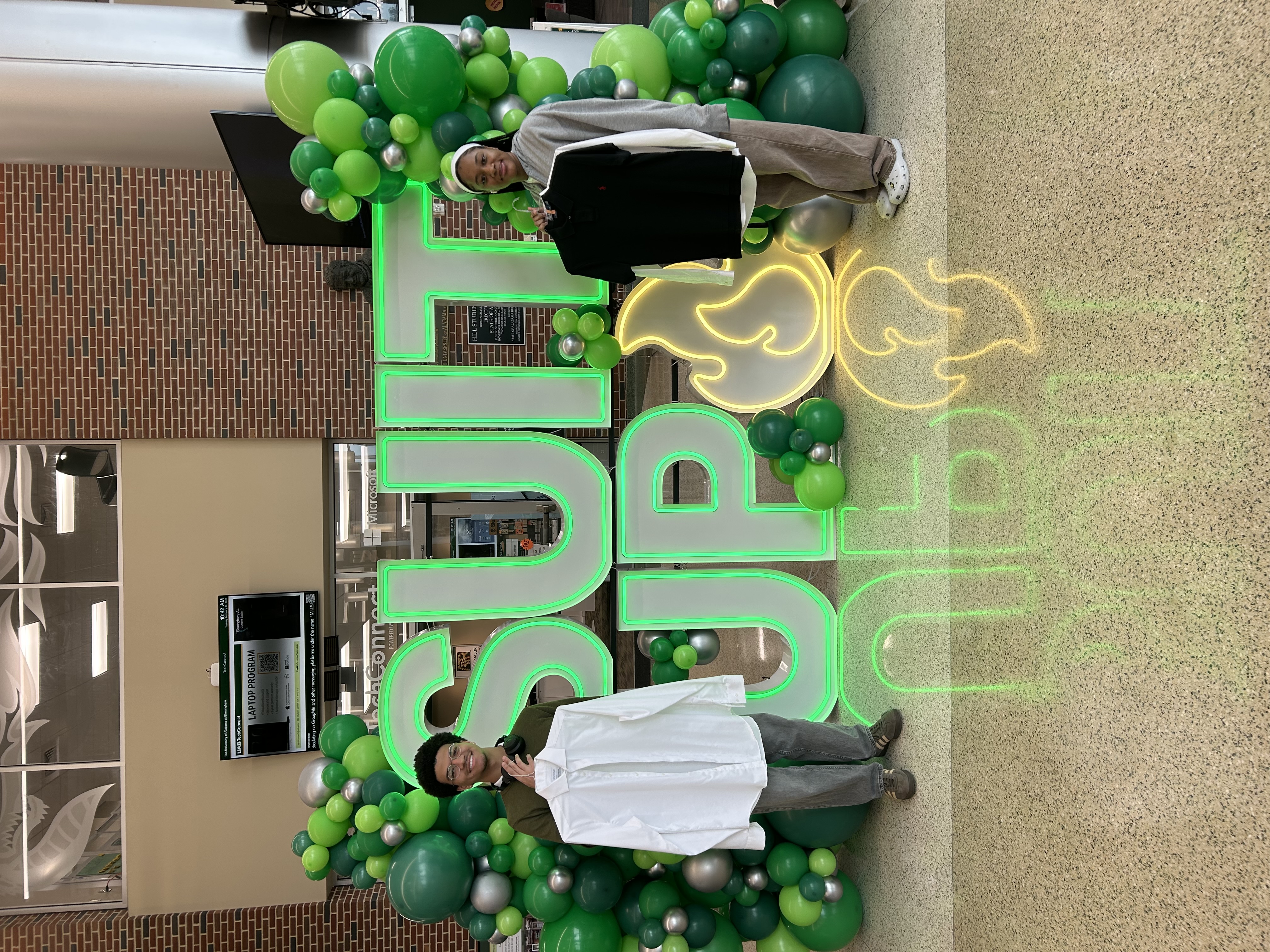 Two UAB students holding professional clothing in front of a lit SUIT UP sign at the Hill Student Center.