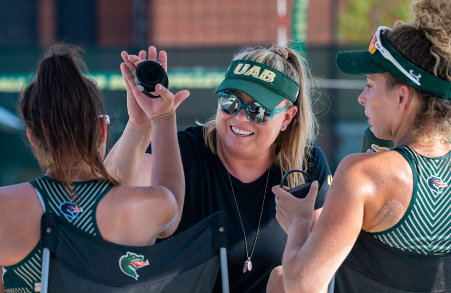 Coach Kyra Iannone meets with two players on the sidelines during a beach volleyball match