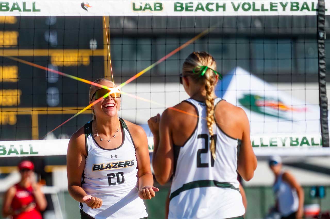 Two smiling beach volleyball players are celebrating during a game at the UAB Beach Volleyball Courts.