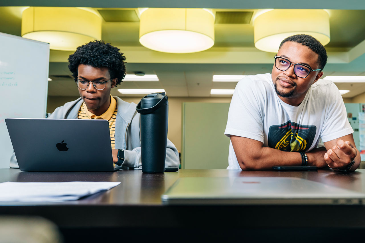 Two students sit at a table with a laptop and water bottle.