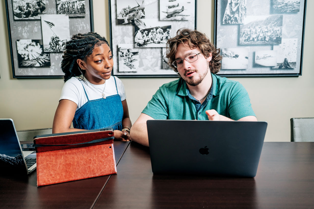 Two students work together at a table with laptops and a tablet.